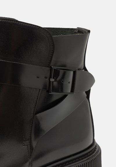 Botină din piele neagră până la gleznă, cu o textură netedă, curea lată cu cataramă și un design conturat pe partea superioară. Talpă plată, texturată.