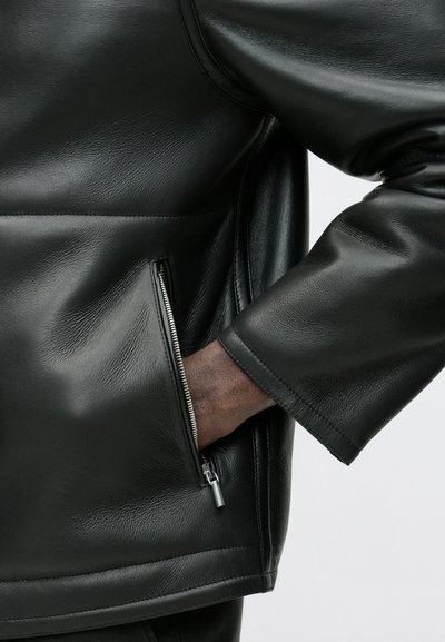 Schwarze Lederjacke mit glatter Textur, seitlicher Reißverschlusstasche und breiten Bündchen für eine entspannte Passform. Klare Linien und minimalistische Details.