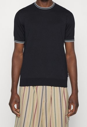 Personne portant un pull noir à manches courtes avec des poignets et un col côtelés rayés gris, associé à un pantalon beige à larges jambes rayé.
