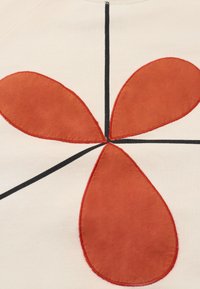 Vêtement crème avec une grande application en forme de goutte rouge, ornée de lignes de couture noires qui se croisent, créant un design géométrique.