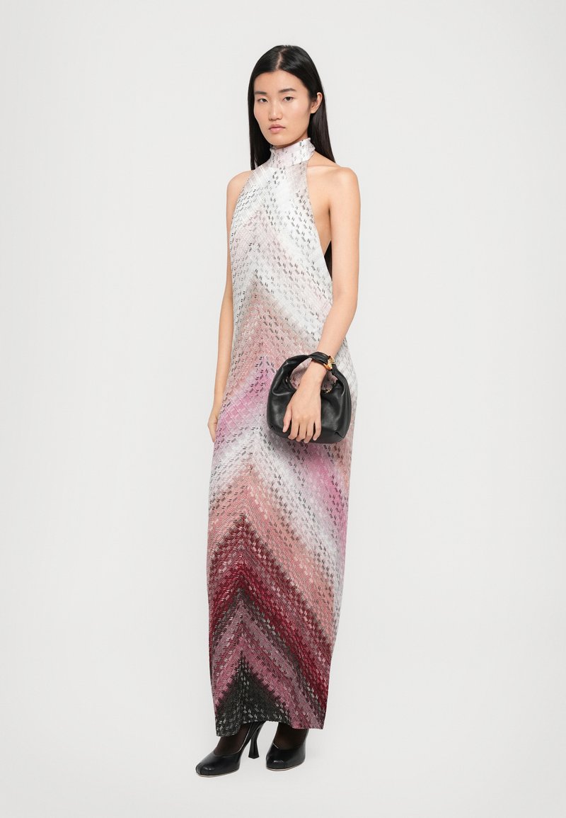 Robe maxi à col halter avec un motif en chevrons dégradés, présentant des nuances de blanc, rose et noir. Complétée par un sac à main noir et des talons.