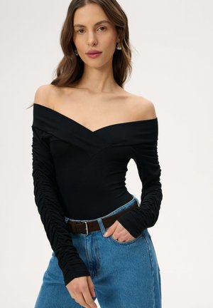 Kobieta w czarnej, marszczonej bluzce z długim rękawem i odkrytymi ramionami, ubrana w niebieskie dżinsy i brązowy pasek, pozująca z jedną dłonią w kieszeni.