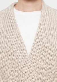 Närbild av en person som bär en beige grovstickad cardigan över en vit ribbad t-shirt med rund hals.