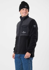 Giacca in pile nera e grigia con chiusura a snap, tasca sul petto e tasche laterali. Presenta un logo sul petto e sulla manica. Abbinata a jeans in denim.