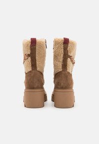 Brązowe zamszowe botki za kostkę z teksturowanym kremowym futrem wewnątrz, czerwonymi akcentami z tyłu i masywną beżową podeszwą. Dekoracyjny pleciony detal z boku.
