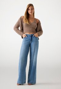Pulover scurt, maro, pufoș, blugi ușori de culoare albastră deschisă cu talie înaltă și picioare largi, cu buzunare frontale, și sandale cu curele. Material neted și croială lejeră.