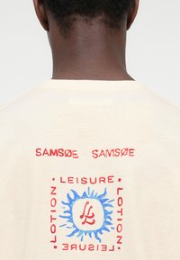 Πίσω όψη ατόμου που φοράει εκρού πουκάμισο με κόκκινο κείμενο "SAMSØE SAMSØE" και μπλε γραφικό ήλιου περιβαλλόμενο από κόκκινο κείμενο "LEISURE ROTION."