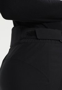 Zwarte broek met een brede tailleband en een klittenbandsluiting. Glad, elastisch materiaal met een getailleerde vorm en subtiele stikseldetails.