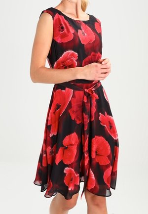 Robe noire sans manches avec de grands imprimés floraux de coquelicots rouges et une ceinture à la taille portée par une personne les mains jointes devant.