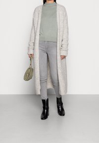 Długi, jasnoszary kardigan wykonany z miękkiego, teksturowanego materiału, w połączeniu z miętowozielonym swetrem, szarymi dżinsami i czarnymi botkami. Trzyma zieloną torbę.
