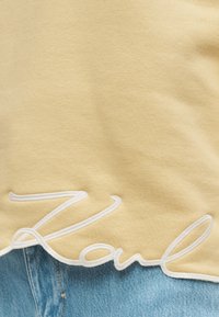 Κίτρινο βαμβακερό μπλουζάκι με λευκή κεντημένη λέξη "Karl" στο τελείωμα. Το τελείωμα είναι ελαφρώς κυρτό, αποκαλύπτοντας τζιν σε ανοιχτό μπλε χρώμα από κάτω.