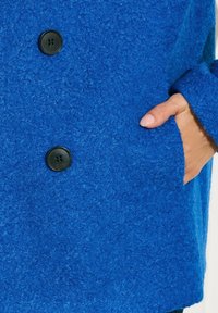 Sininen villakacos, jossa on teksturoitu pinta. Varustettu mustalla napilla ja sivutaskulla. Suunnittelussa on rento leikkaus ja pitkät hihat.