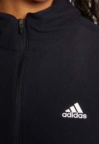 Gros plan sur une personne portant une veste zippée noire Adidas avec le logo blanc Adidas sur la poitrine.