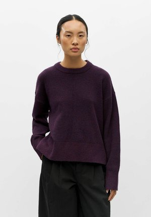 Femme aux cheveux foncés attachés, portant un pull violet foncé et un pantalon noir sur un fond clair uni.