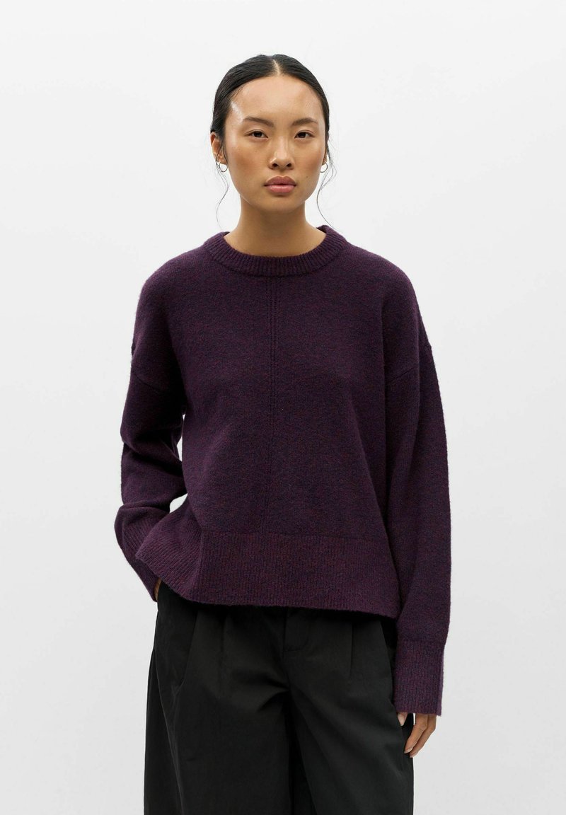 Femme aux cheveux foncés attachés, portant un pull violet foncé et un pantalon noir sur un fond clair uni.
