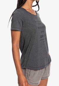 Camiseta gris de manga corta con un corte relajado, que presenta un diseño gráfico de texto en la parte delantera y un escote redondeado. Textura de tela suave.