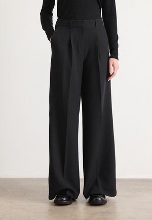 Wide black trousers with a smooth texture, pleats at the front, and side pockets. Worn with a fitted black top and black shoes.