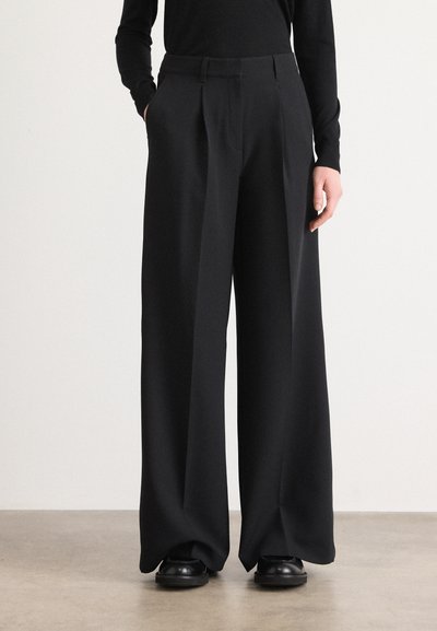 Pantalons noirs larges avec une texture lisse, plis à l'avant et poches latérales. Portés avec un haut noir ajusté et des chaussures noires.