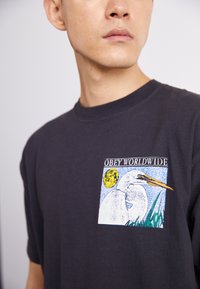 Černé bavlněné tričko s grafickým potiskem volavky v noci a textem "OBEY WORLDWIDE" v barevném rámcovém designu na hrudi.