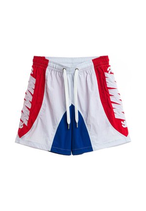 Joven de pie mirando hacia adelante con camiseta roja "DOG'S PARTY", pantalones cortos blancos y azules, calcetines blancos y zapatos beige texturizados sobre un fondo blanco liso.