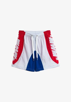 Joven de pie mirando hacia adelante con camiseta roja "DOG'S PARTY", pantalones cortos blancos y azules, calcetines blancos y zapatos beige texturizados sobre un fondo blanco liso.