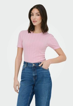 Woman with dark hair wearing a light pink short-sleeve ribbed top and high-waisted blue jeans, hand in pocket, neutral background.