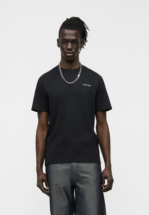 Νεαρός με σκουρόχρωμο δέρμα και dreadlocks, φορώντας μαύρο t-shirt Calvin Klein, ασημένιο αλυσίδα λαιμού και γκρι τζιν, στέκεται μπροστά σε λευκό φόντο.