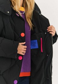 Czarny pikowany płaszcz z kolorowymi guzikami (różowy, pomarańczowy, fioletowy) oraz kieszenią na zamek z niebieską metką z napisem. Strukturalny kołnierz i materiał.