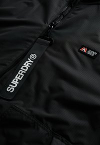 Zwarte zip-up jas van lichtgewicht stof met een gestructureerd oppervlak. Details zijn onder meer een logo-tag en een kleine merkembleem op de borst.