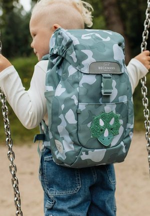 Zaino verde mimetico con una patch texturizzata di un dinosauro, spallacci regolabili, chiusura con patta superiore e una tasca frontale. Adatto per bambini.