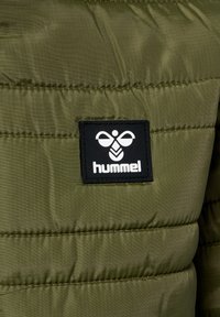 Grön puffjacka med en slät, glänsande textur och horisontell sömnad. Har en svart rektangulär logotypskylt med en vit design.