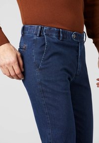 Homme portant un jean slim bleu foncé et un haut marron à manches longues, debout avec les mains près des hanches sur un fond uni.