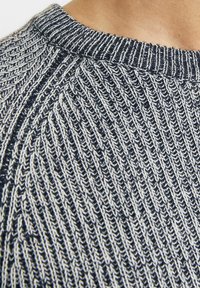 Pulover tricotat texturat, cu un scheme de culori bleumarin și crem, având un decolteu rotund cu dantelă și modele de cusut diagonale.