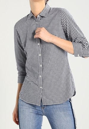 Person wearing a black and white checkered button-up shirt with three-quarter sleeves and blue jeans with a dark side stripe.