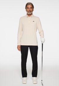 Beige langärmliges Polohemd mit schwarzem Logo, kombiniert mit schwarzen Hosen und weißen Sneakers. Das Model hält einen Golfschläger und trägt einen weißen Golfhandschuh.