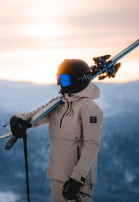 Giacca da sci beige con dettagli e cerniere nere, casco nero con visiera blu riflettente, che tiene sci e bastoncini da sci. Sfondo montano.