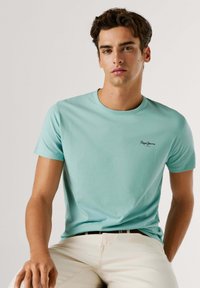 Jaunas vyras trumpais tamsiais plaukais, dėvintis šviesiai turkio spalvos Pepe Jeans marškinėlį ir baltas kelnes, sėdi priešais paprastą šviesią foną.