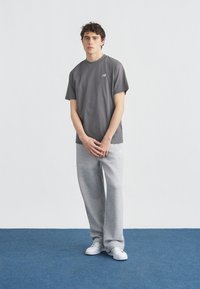 Νέος άνδρας στέκεται σε μπλε χαλί φορώντας ανοιχτογριζό φανελάκι New Balance, ανοιχτογριζό φόρμα και λευκά αθλητικά παπούτσια μπροστά σε άσπρο τοίχο.