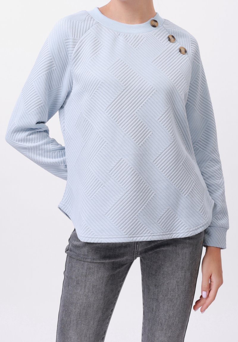 Pull bleu clair texturé avec des motifs en côtes diagonales et trois boutons marron sur l'épaule gauche, porté avec un jean gris délavé.