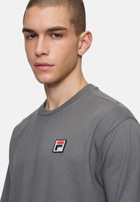 Grå bomulds t-shirt med rund hals, med et lille broderet logo i rød, hvid og blå på venstre bryst.