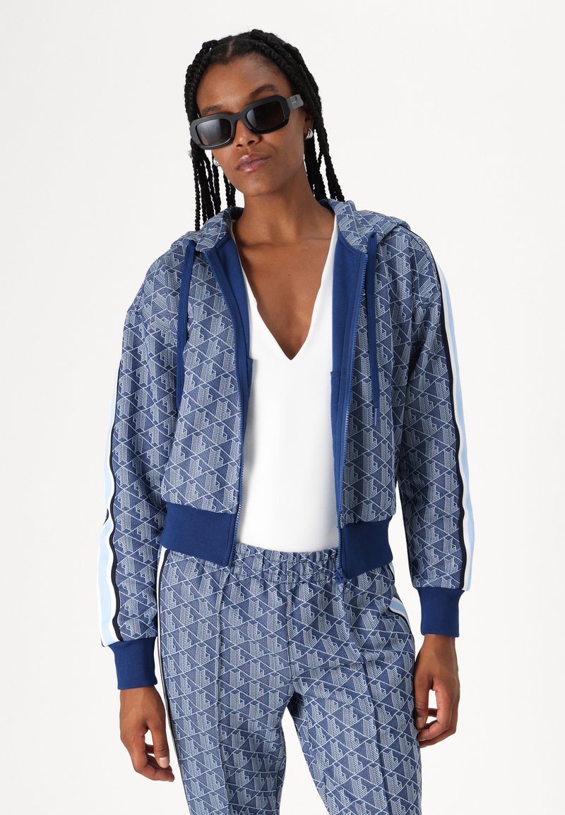 Felpa con cappuccio blu a fantasia abbinata a pantaloni, caratterizzata da un design geometrico, cappuccio con cordino e polsini a coste. Modello indossa occhiali da sole neri.