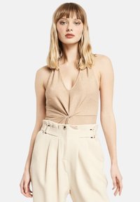 Top halter neck dorato metallico con dettagli frontali annodati, abbinato a pantaloni crema a vita alta con pince e passanti per cintura.