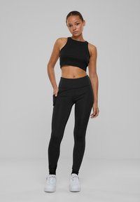 Top cropped nero e leggings a vita alta, entrambi con una superficie liscia. I leggings sono dotati di tasche laterali, abbinati a sneakers bianche.