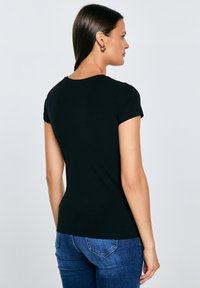 Vrouw met lang donker haar, gouden oorringen in de vorm van ringen, een zwart nauwsluitend T-shirt met korte mouwen en een blauwe spijkerbroek, gezien vanaf de achterkant.