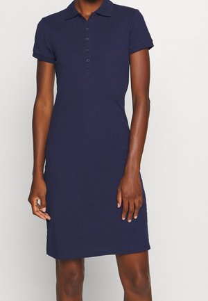 Mujer con un vestido tipo polo azul marino de manga corta con tapeta abotonada, de pie frente a un fondo liso y claro.