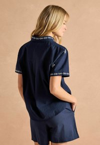 Cămașă bleumarin cu mânecă scurtă, cu broderie albă pe guler și pe mâneci, realizată dintr-un material texturat. Are un croi lejer și buzunare laterale.