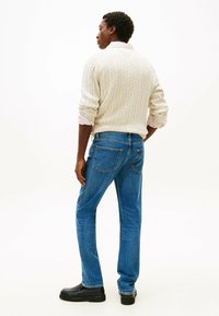 Cremfarbener Kabelstrickpullover, blaue Jeans mit Gesäßtaschen und schwarze Schuhe. Modell von hinten gezeigt, mit sichtbaren strukturierten Details.