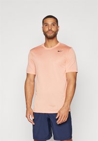 Vaaleanpunainen Nike lyhythihainen urheilupaita, jossa on pyöreä kaula-aukko ja musta logo rinnassa. Pehmeä, sileä kangas. Yhdistetty tummansinisiin shortsihousuihin.