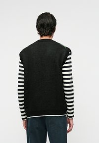 Schwarze gestrickte Weste mit weiten Armausschnitten, getragen über einem schwarz-weißen, gestreiften Langarmshirt; mit grünen gestickten Akzenten an den Schultern.