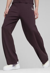 Donkerpaarse geribde wijde broek met een elastische tailleband en een kleine gouden logoaccent. Draagt met witte sneakers met paarse details.